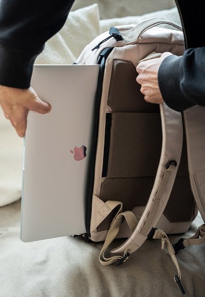 Mochila beige claro con cremalleras negras, que cuenta con un compartimento acolchado para un portátil. Se está insertando un portátil plateado con un logo rosa.