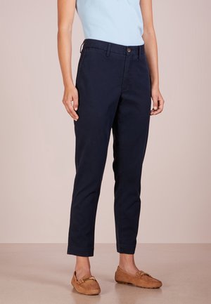 Kvinde iført marineblå taperede bukser, lyseblå ærmeløs top og tanfarvede loafers, stående mod en ensfarvet beige baggrund.