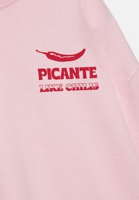 Rózsaszín pulóver piros felirattal, amelyen a „PICANTE” szöveg olvasható egy chili paprika grafika felett, alatta pedig vastag, laza stílusú betűkkel a „LIKE CHILIS” felirat.