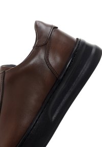 Brown leather slip-on shoe with black thick sole, visible stitching, and smooth finish, angled to show side profile and heel support.