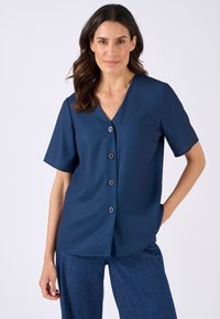 Top blu navy a maniche corte con scollatura a V, sei bottoni sul davanti e un tessuto liscio. Vestibilità su misura con dettagli di sagomatura discreti.