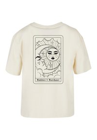 Krémové tričko s černým grafikou slunce a měsíce s obličejem, obklopené mraky a hvězdami, v rámu podobném kartě.
