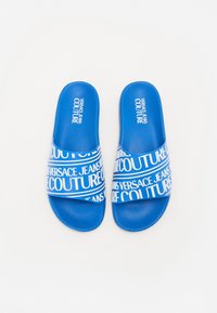 Plave pjenaste natikače s teksturiranom površinom. Bijeli logo "Versace Jeans Couture" otisnut na trakama, s uzorkom koji se ponavlja.