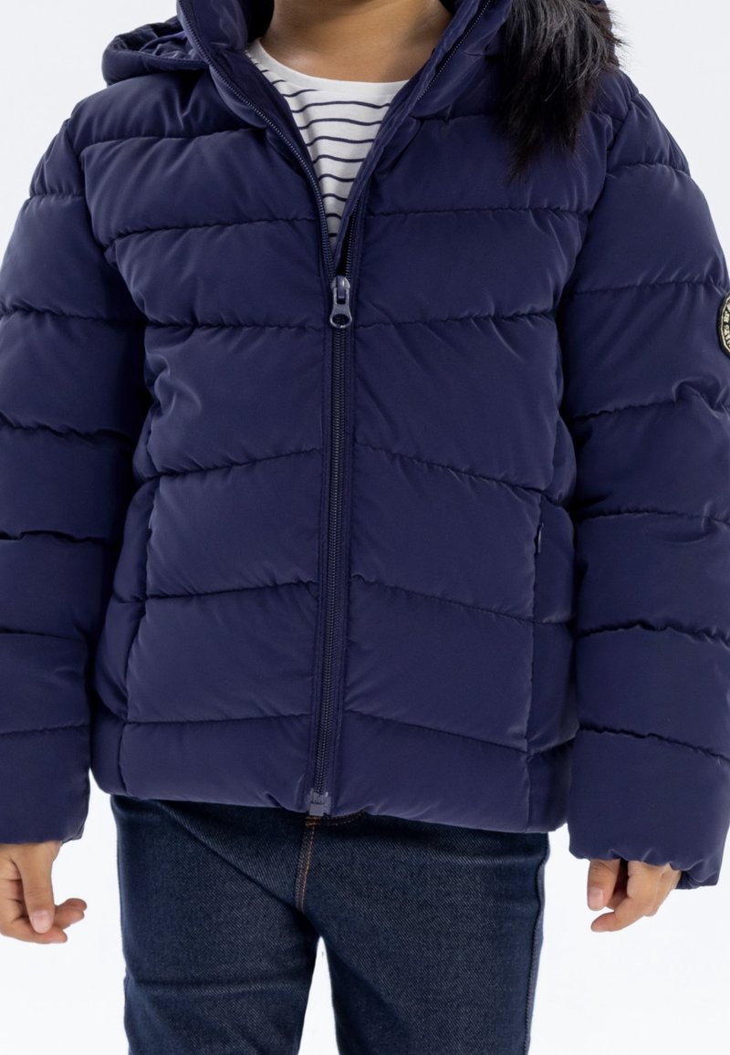 Enfant portant une doudoune bleue marine zippée sur un t-shirt rayé et un jean foncé, debout les mains détendues le long du corps.