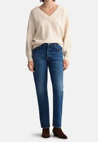 Cremfarbener, gerippter Strickpullover mit breitem V-Ausschnitt, kombiniert mit dunkelblauben, geraden Jeans und braunen Ankle Boots.