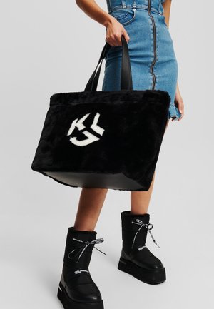 Sac cabas en fausse fourrure noire avec un logo blanc, doté de poignées en cuir noir et d'un design spacieux, associé à des bottes plateformes noires.