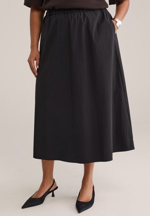 Jupe midi noire avec taille élastique portée avec des escarpins slingbacks noirs à bouts pointus sur un mannequin devant un fond neutre.