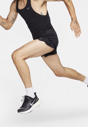Läufer in schwarzer Sportkleidung und Nike-Sneakern sprintet vor weißem Hintergrund, sichtbare Tattoos an Armen und Beinen.