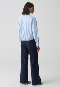 Blusa a righe blu chiaro con maniche lunghe e orlo elastico. Indossata con pantaloni ampi in denim scuro e scarpe chiare. Sfondo neutro.