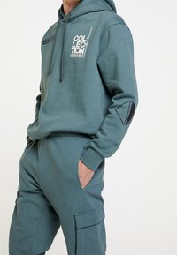 Set van een hoodie en cargo-broek in tealkleur. Voorzien van trekkoorden, logo-opdruk op de borst en zakken op de broek met een gestructureerde afwerking.