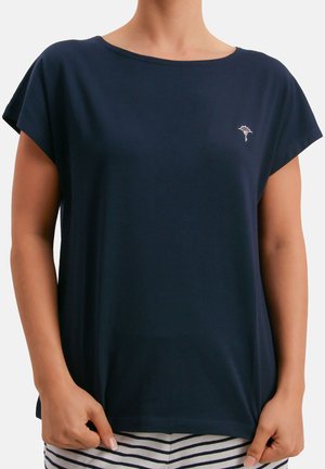 Persona vistiendo una camiseta azul marino de manga corta y corte holgado con un pequeño logo floral blanco en el pecho, combinada con pantalones a rayas blancas y azules marinas.