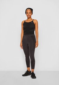 Top desportivo preto com detalhes franzidos, combinado com leggings pretas que apresentam um padrão texturizado. Modelo usa ténis pretos.