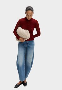 Maglione a collo alto rosso con texture, jeans blu larghi e scarpe nere basse. Tiene una borsa beige a pieghe. Outfit semplice e casual.