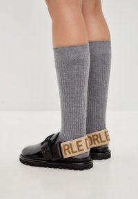 Sandalias de charol negro con suela gruesa, que presentan una tira en la muñeca beige con letras en relieve. Medias altas de color gris con canalé.