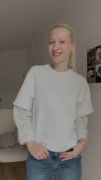 Weißer oversized Pullover mit gerippten langen Ärmeln, kombiniert mit blauen Jeans. Weicher Stoff und lockere Passform, minimalistisches Design.