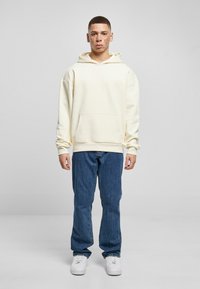 Sudadera con capucha en color crema y bolsillo frontal, combinada con vaqueros rectos azules y zapatillas blancas. El modelo se encuentra de pie frente a un fondo gris.