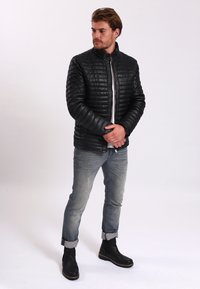 Svart vadderad jacka med hög krage, dragkedjestängning, tillsammans med ljusblå jeans och svarta ankelskor, stående på en vit bakgrund.