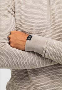 Helle beige Sweatshirt mit einem strukturierten Wabenmuster. Verfügt über einen halben Reißverschluss am Kragen und enganliegende Bündchen mit einem Markenlabel am Handgelenk.