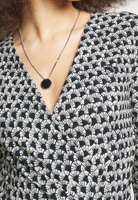 Schwarzes und weißes gemustertes Wickeloberteil mit V-Ausschnitt. Silberne Kettenhalskette mit einem runden schwarzen Anhänger. Auffälliges fächerförmiges Design.