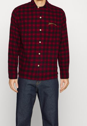 Camisa de cuadros roja y negra hecha de franela, con cuello, cierre de botones y un logo bordado en dorado en el pecho.