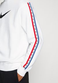 Vit sweatshirt med svart Nike-logotyp, med röda och blå sidostripes som har ett upprepat Nike-textmönster. Mjukt, gulligt material.