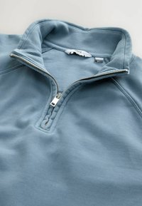 Lichtblauwe quarter-zip pullover van zacht stof, met een geribde kraag en een metalen rits met een zilveren trekker.