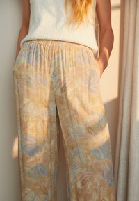 Kvinna klädd i lösa blommiga byxor i pastellfärger och en ärmlös vit top med struktur, stående bredvid en gardin i mjukt ljus.