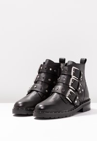 Černé kožené kotníkové boty s třemi nastavitelnými pásky se stříbrnými přezkami a hřebíky, plochou podrážkou a mírně texturovaným povrchem.