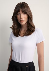 Młoda kobieta o średniej długości brązowych włosach z grzywką, ubrana w białą koszulkę i czarną plisowaną spódnicę, na jednolitym jasnym tle.