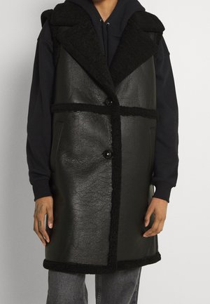 Person iført en lang sort lædervest foret med fåreskind over en sort hættetrøje og grå jeans, stående mod en neutral baggrund.