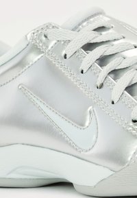 Zapatilla plateada con un acabado brillante, cordones blancos y un sutil logo de Nike en relieve. Suela texturizada con patrón de goma visible.