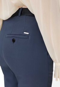 Pantalon bleu marine sur mesure avec poche arrière à bouton et petite étiquette de marque métallique, porté avec un chemisier crème à manches longues avec poignets froncés.