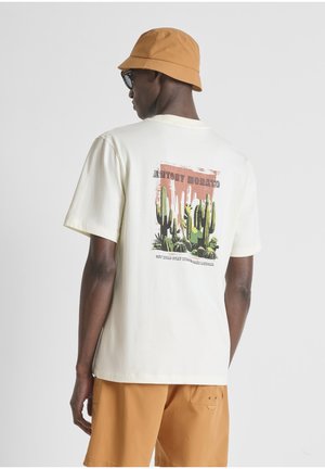 Man draagt een wit T-shirt met cactusafbeelding en tekst op de achterkant, beige bucket hat, zonnebril en oranje korte broek, met de rug naar de camera.