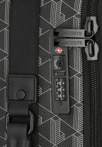 Valise noire avec motif géométrique, deux tirettes de fermeture éclair Lacoste et un cadenas à combinaison approuvé par la TSA réglé sur 0-0-0.