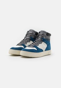 Vysoké tenisky s modrou semišovou částí, šedou síťovinou a bílými akcenty. Silná krémová gumová podrážka se šedými tkaničkami a detailem loga na jazyku.