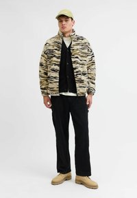 Chaqueta de forro polar con un patrón de rayas de tigre en beige y negro, superpuesta sobre un cárdigan negro y una camiseta blanca, combinada con pantalones negros y zapatos beige.
