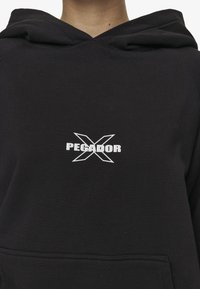 Sudadera negra de algodón, con un bolsillo frontal. Gráfico "PEGADOR" con un logo de "X" en blanco en el pecho. Textura suave.