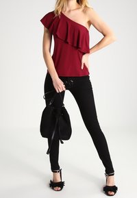 Top burdeos de un solo hombro con detalle de volantes, combinado con jeans ajustados negros y un bolso de cubo negro. Sandalias negras con acentos de volantes y hardware circular.