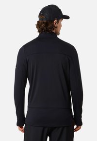 Zwarte sportieve longsleeve met een opstaande kraag, geweven stof en gestikte details op de schouders en zijkanten, gedragen met een zwarte pet.