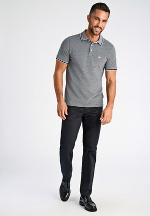 Homme marchant, portant un polo gris à manches courtes, un pantalon noir et des chaussures habillées noires et brillantes, souriant devant un fond clair uni.