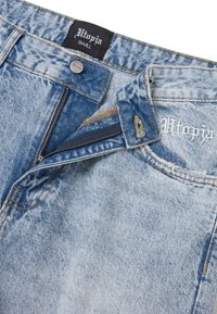 Vaqueros de mezclilla azul claro con cremallera, con una superficie texturizada, costuras en contraste y una etiqueta que dice "Utopía" en blanco.