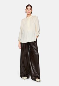 Cremfarbene Bluse mit langen Ärmeln, Stehkragen und Knopfdetails, kombiniert mit hoch taillierten, weiten, glänzenden schwarzen Lederhosen.