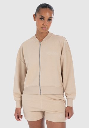 Beige huppari vetoketjulla, jossa on rento malli, ribbaidut hihansuut ja vyötärö. Vasemmassa rinnassa brodeerattu logo. Yhdistetty beigeihin shortseihin.