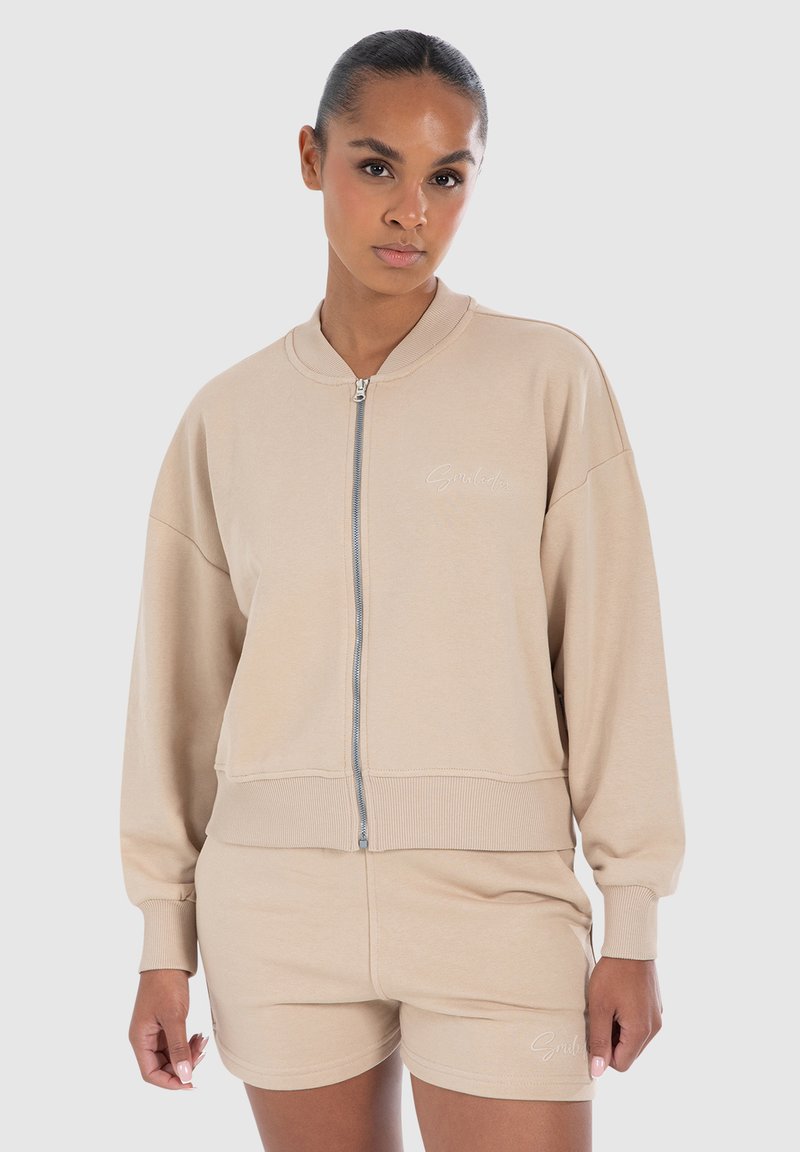 Sweat-shirt zippé beige avec une coupe décontractée, poignets et taille côtelés. Présente un logo brodé sur la poitrine gauche. Associé à un short beige.