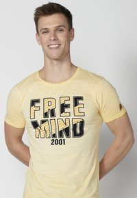 Dzeltena kreklu ar apaļu kakla izgriezumu, kurā ir bolda melna un dzeltena svītra teksts "FREE MIND 2001". Mīksts audums un pieguļošs dizains.