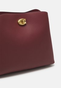 Sac à main en cuir bordeaux avec une texture lisse et un fermoir en métal doré. Le design est structuré avec une large ouverture en haut.