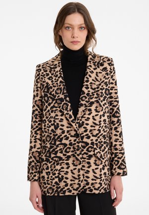 Woman wearing a leopard print blazer over a black turtleneck and black pants, standing against a plain white background.