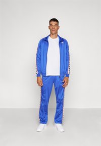 Blauwe trainingsoutfit van gladde stof. De jas heeft een rits en gestreepte zijkanten; de broek heeft bijpassende strepen. Het model draagt een wit t-shirt en schoenen.