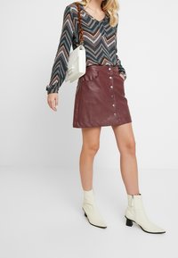 Mini-jupe en cuir bordeaux avec fermeture à boutons sur le devant et poches, associée à un haut à manches longues à motifs et des bottines blanches.