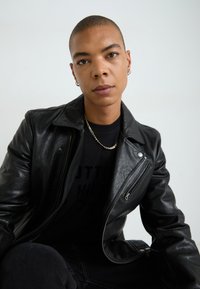 Must musta bikiersinine jakk, millel on tõmblukkude detailid, kantud musta T-särgi peal. Tuntavad rõhutused hõbedasest ketist kaelakee ja kõrvarõngad.